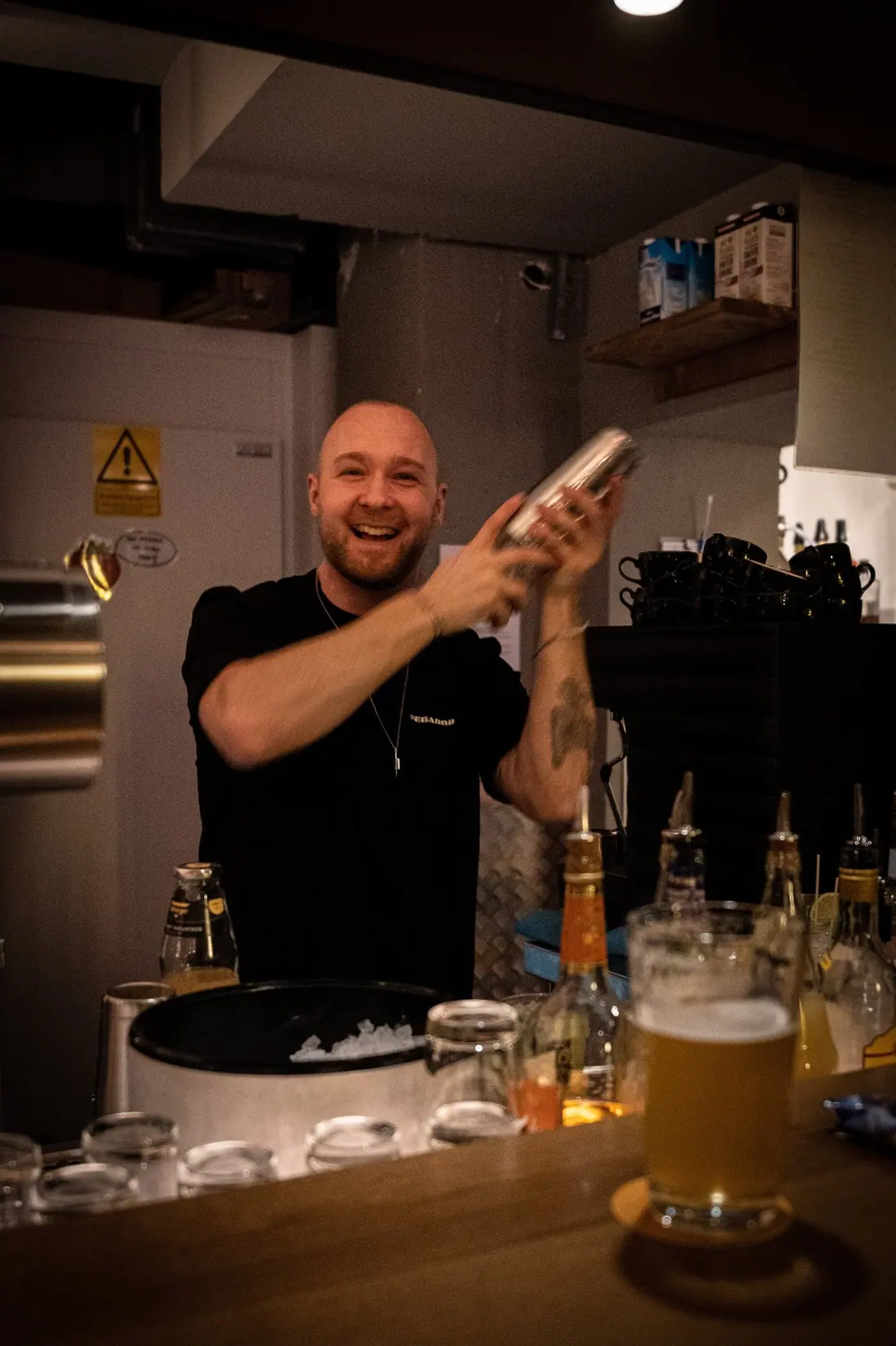 Ein Barkeeper schüttelt lachend einen Cocktail, vor ihm ein frisch gezapftes Bier
